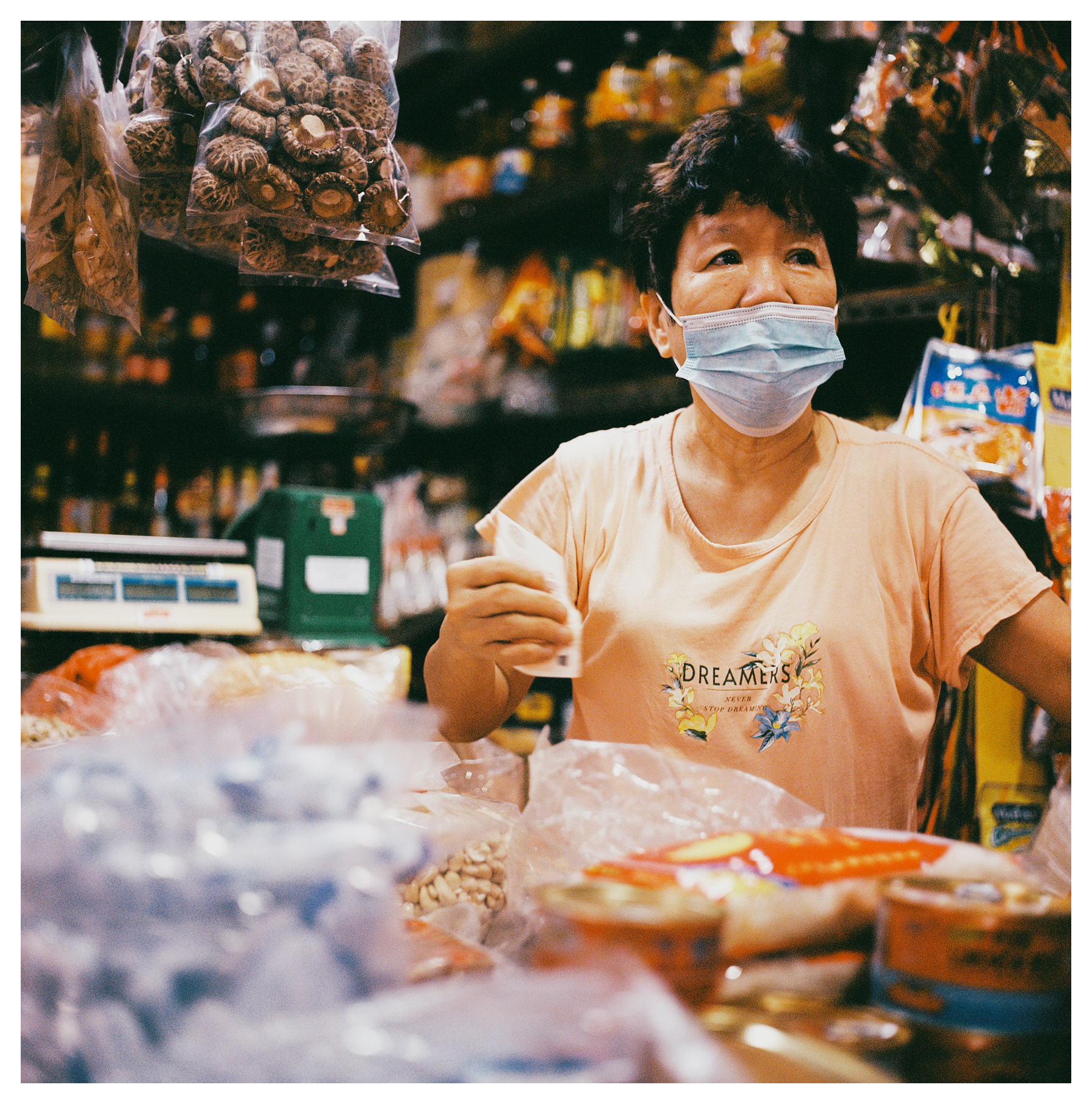 A scan of a medium format square color photo of an older Chinese lady wearing a face mask and tending a store in a market in Singapore