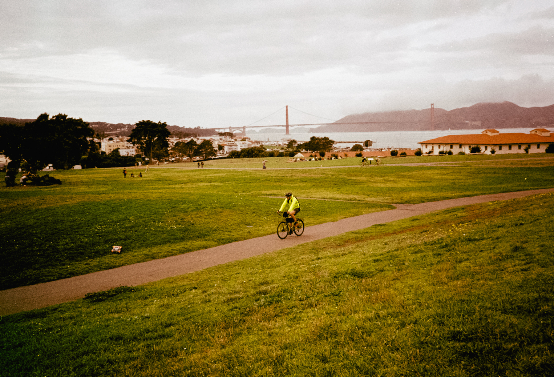a scan of a color photo of a cyclist biking through fort mason in san francisco with a fluorescent jacket. in the background, the golden gate bridge is slightly obscured
