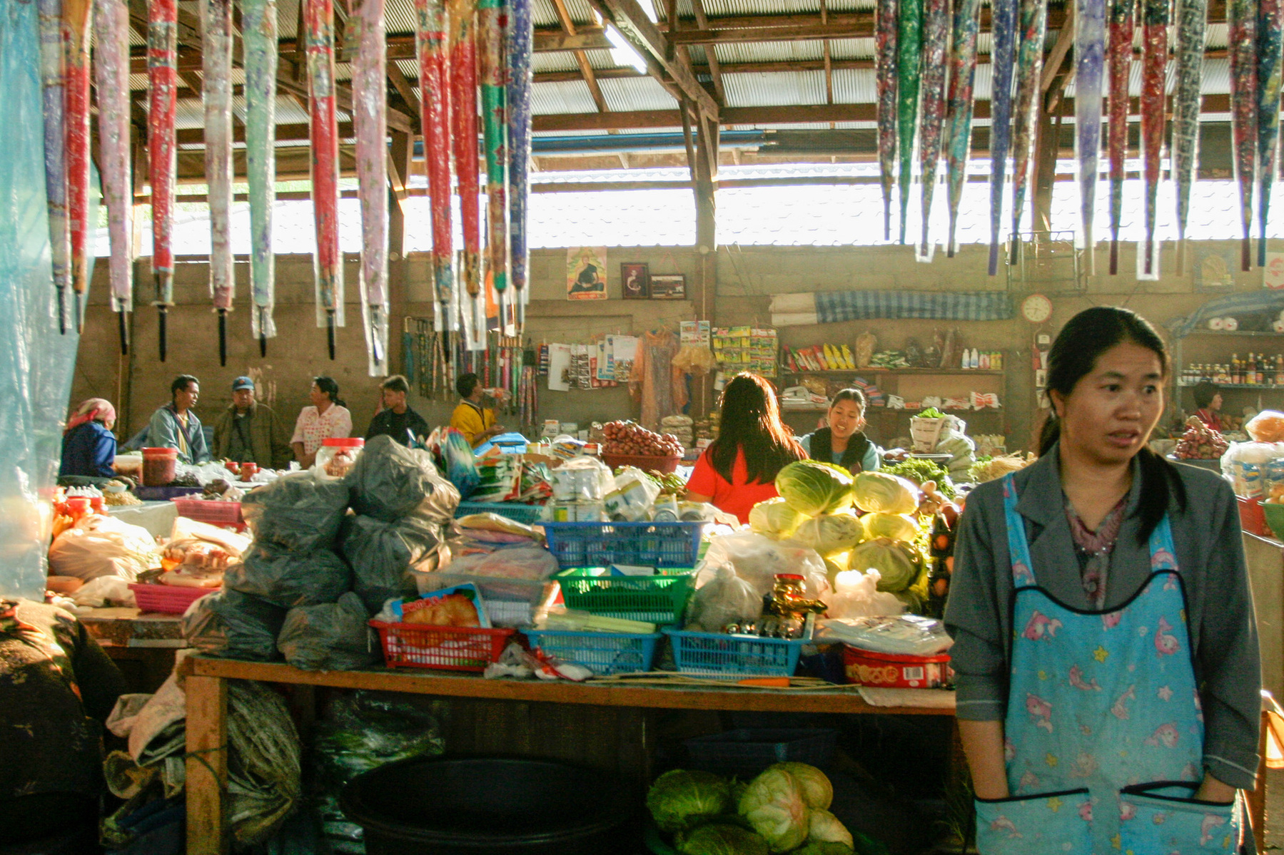 A woman in a rural Thai market standing in front of a camera while others do business in the background