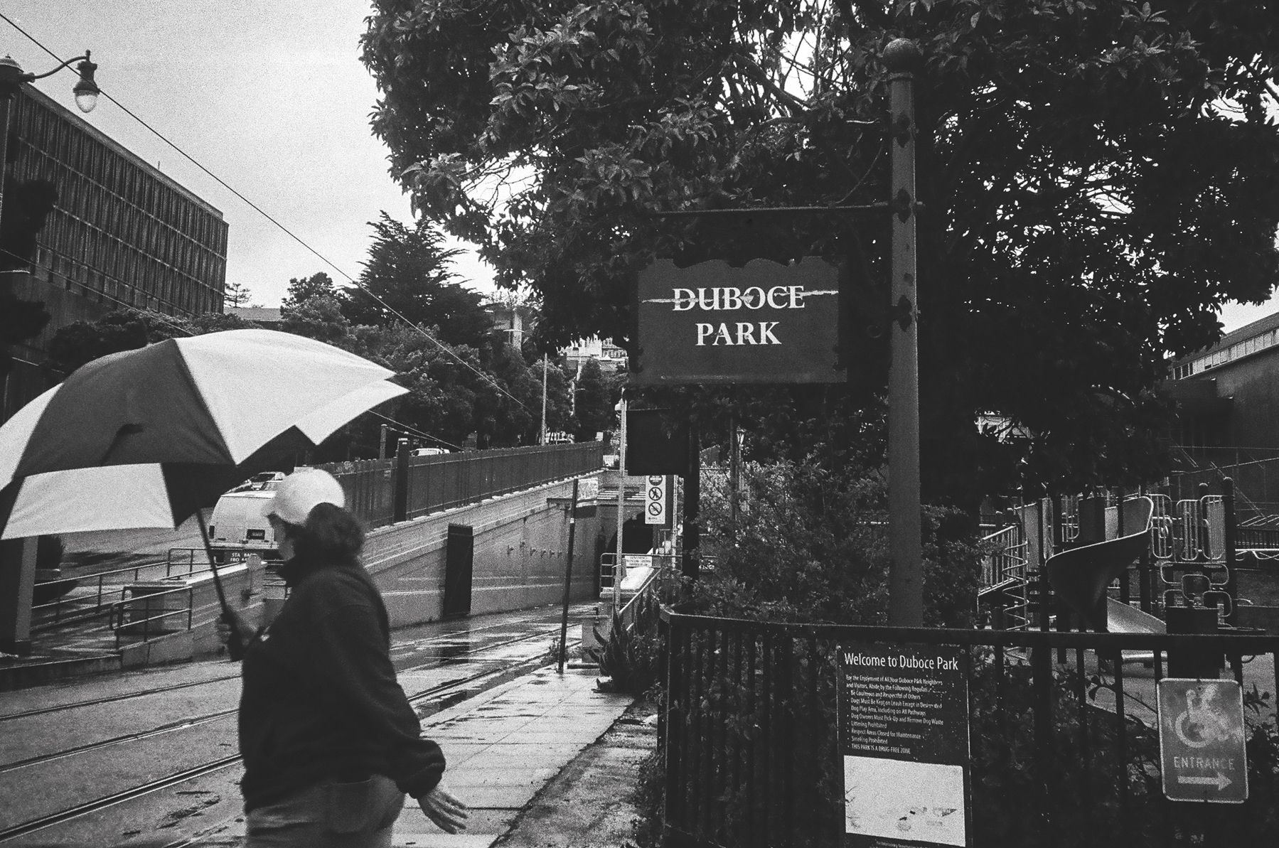a scan of a black and white photograph of woman holding an umbrella standing at Duboce Park waiting for a train in the rain