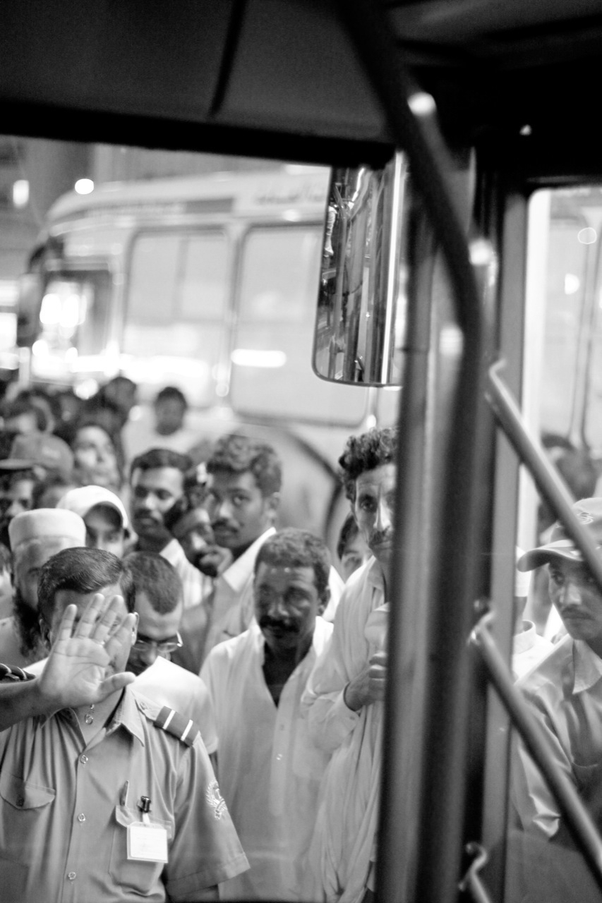 a digital black and white photograph of some South Asian workers waiting to board a bus in Dubai