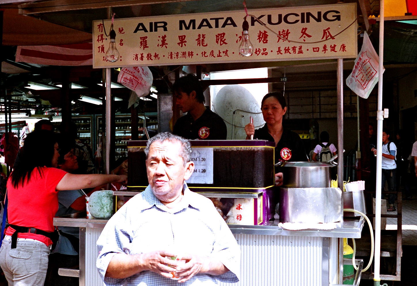 A color photograph of a person standing in front of a Malaysian beverage stall with signs on it