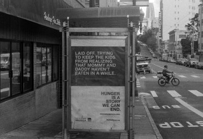 a scan of a black and white photo showing a bus stop with a sign, laid off and trying to keep the kids from realizing that mommy and daddy haven't eaten in a while. behind, a gig worker zooms past on an ebike, and in the far background, the salesforce tower looms large
