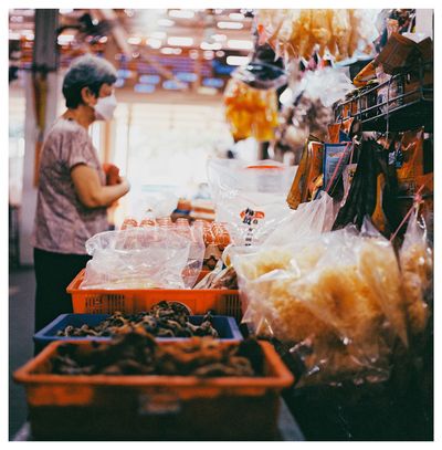 A scan of a medium format square color photo of an older Chinese lady wearing a face mask standing at a stall in Singapore looking at dried goods to purchase