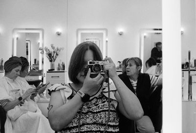 A person photographs themselves in a salon mirror holding a film camera, black and white