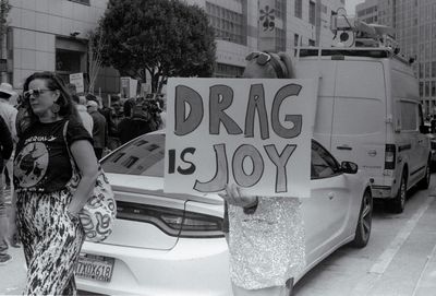 a scan of a black and white photo showing someone holding a sign that says Drag is Joy