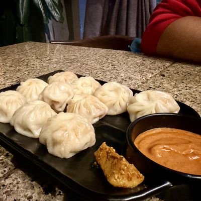 Plump steamed momos on a black plate beside a bowl of orange dipping sauce
