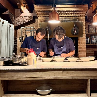 Two cooks plating food beneath copper pendant lights in a kitchen framed by brickwork and carved wood