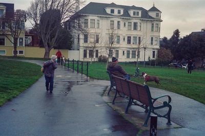 a scan of a color photograph of an old lady walking through a park looking at people who are playing with their dogs in a dog park