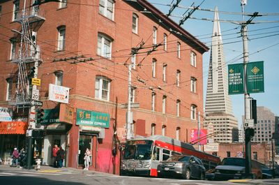 a color photo of the border of Chinatown San Francisco and the Financial District with Chinese shops in the foreground and TransAmerica pyramid in the background, a Muni bus at the bus stop at an intersection, people standing around or crossing the street