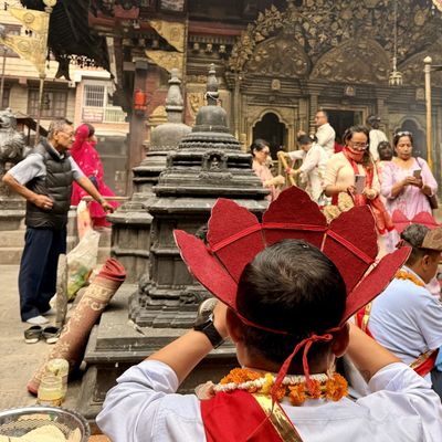 A person wearing a red headdress and marigold garland faces a stone shrine, surrounded by people in an ornate temple courtyard