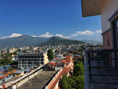 Snow-covered mountains rise behind green hills and city rooftops, viewed from a balcony beneath a clear blue sky